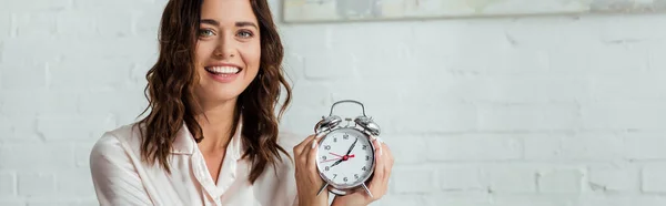 Panoramic shot of attractive woman smiling and holding alarm clock at morning — Stock Photo