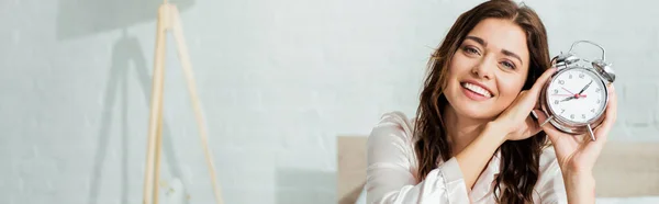 Panoramic shot of attractive woman smiling and holding alarm clock at morning — Stock Photo