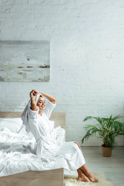 Mujer atractiva en albornoz y toalla sonriendo y sentada en la cama por la mañana - foto de stock