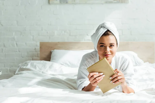 Séduisante femme en serviette et peignoir tenant livre le matin — Photo de stock