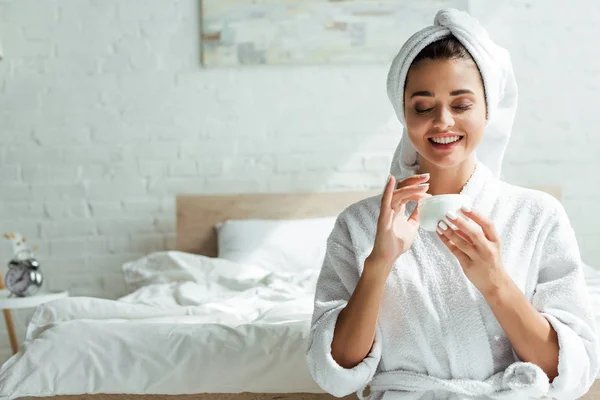 Mulher atraente em roupão de banho e toalha sorrindo e segurando creme cosmético de manhã — Fotografia de Stock