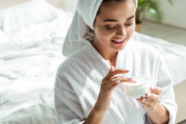Attractive woman in bathrobe and towel smiling and holding cosmetic cream at morning — Stock Photo