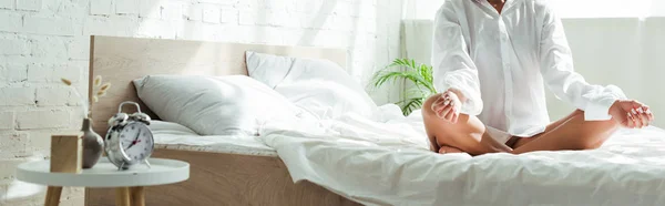 Panoramic shot of woman in white shirt sitting on bed in lotus pose at morning — Stock Photo
