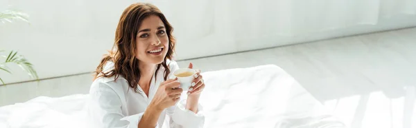 Plan panoramique de la femme attrayante en chemise blanche souriant et tenant tasse le matin — Photo de stock