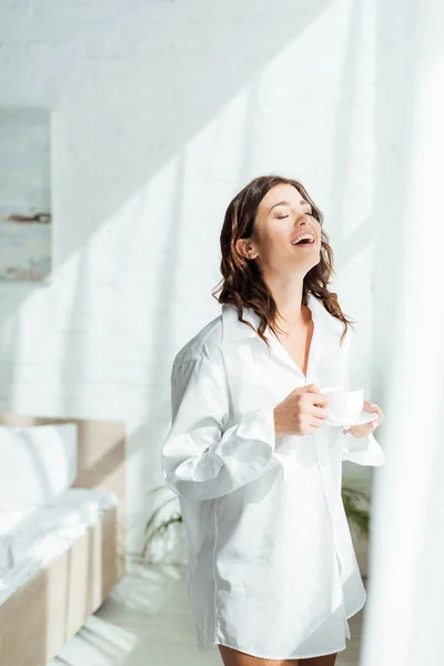 Atractiva mujer en camisa blanca sonriendo y sosteniendo la taza por la mañana - foto de stock
