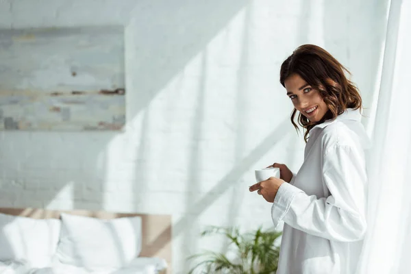 Atractiva mujer en camisa blanca sonriendo y sosteniendo la taza por la mañana - foto de stock