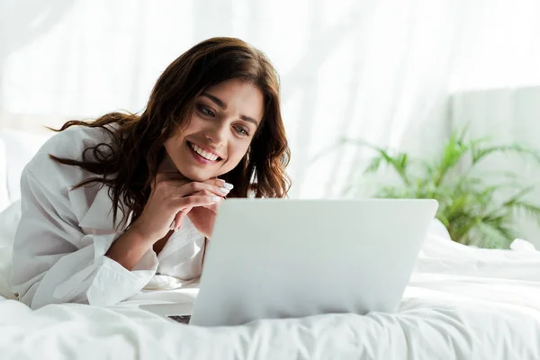 Atractiva mujer en camisa blanca usando portátil y sonriendo por la mañana - foto de stock