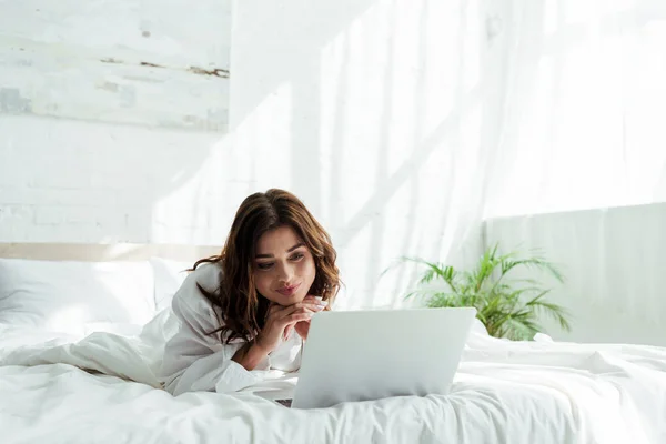 Mujer sonriente en camisa mirando el ordenador portátil por la mañana - foto de stock