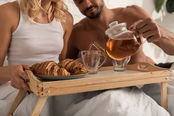 Vista cortada de jovem casal com chá e croissants na bandeja na cama pela manhã — Fotografia de Stock