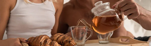 Cropped view of man pouring tea into cups on tray with croissants in bed — Stock Photo