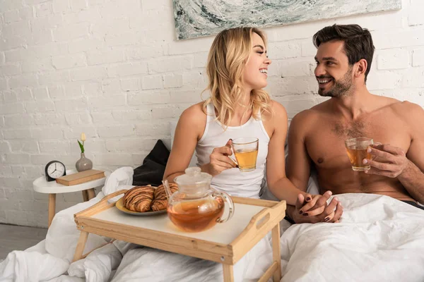 Feliz pareja tomando té y croissants para el desayuno en la cama por la mañana - foto de stock