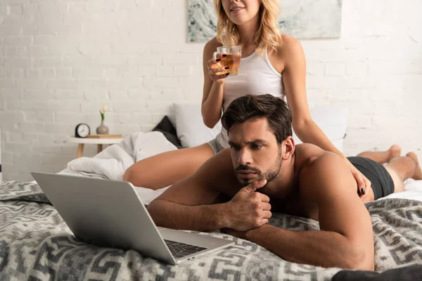 Pensive man using laptop in bed with girlfriend drinking tea behind — Stock Photo