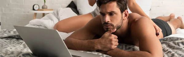 Красивий вдумливий чоловік дивиться на ноутбук в ліжку з дівчиною позаду — стокове фото