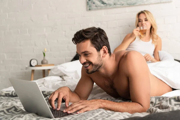 Handsome man using laptop in bed with girlfriend drinking tea behind — Stock Photo