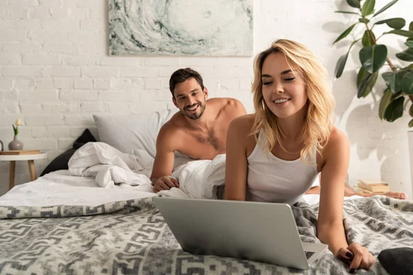 Chica atractiva usando el ordenador portátil en la cama con el novio sonriente detrás - foto de stock