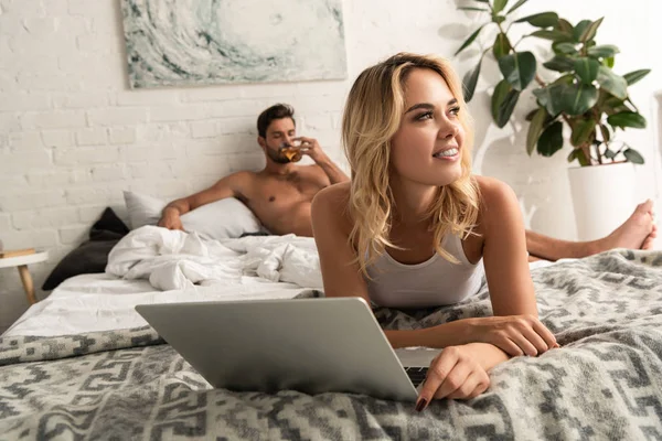 Beautiful dreamy woman using laptop in bed with boyfriend drinking tea behind — Stock Photo