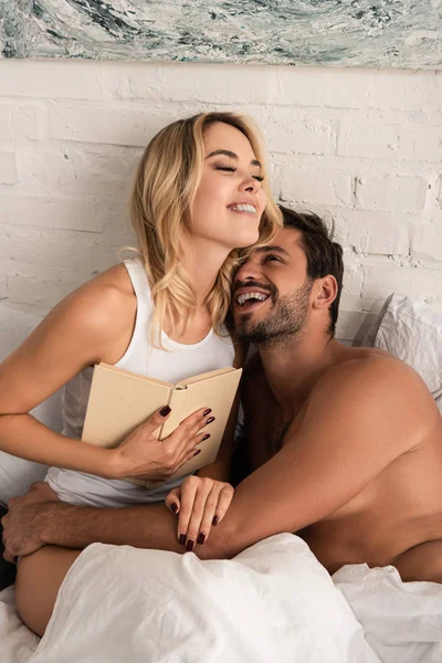 Sonriente pareja abrazando y leyendo libro juntos en la cama por la mañana - foto de stock