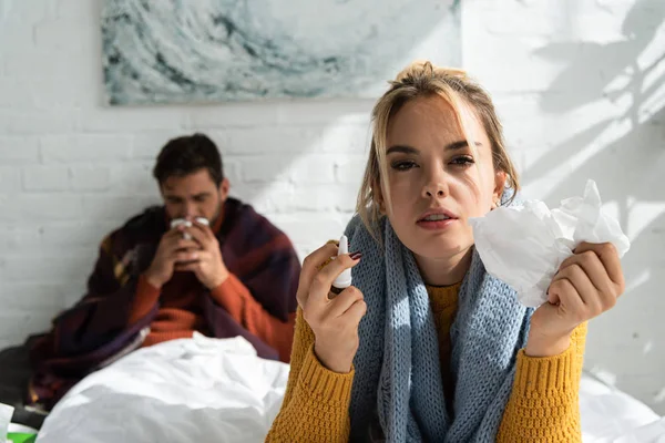 Femme malade tenant pulvérisation nasale et serviettes dans la chambre avec l'homme derrière — Photo de stock