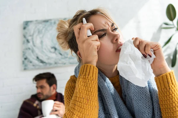 Femme malade avec mal de tête tenant pulvérisation nasale et serviettes dans la chambre avec l'homme derrière — Photo de stock