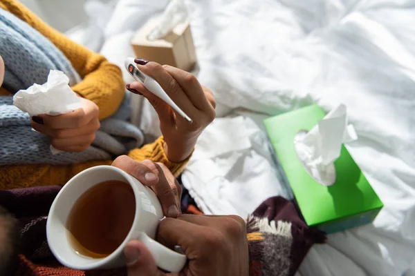 Vista cortada de casal doente segurando termômetro e bebida quente na cama com guardanapos — Fotografia de Stock