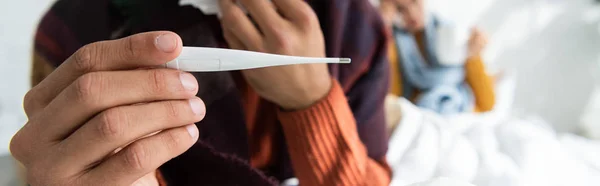 Cropped view of sick man with fever holding thermometer in bedroom with woman behind — Stock Photo