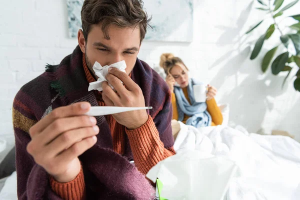 Hombre enfermo con fiebre sosteniendo termómetro y servilleta en el dormitorio con la mujer detrás - foto de stock