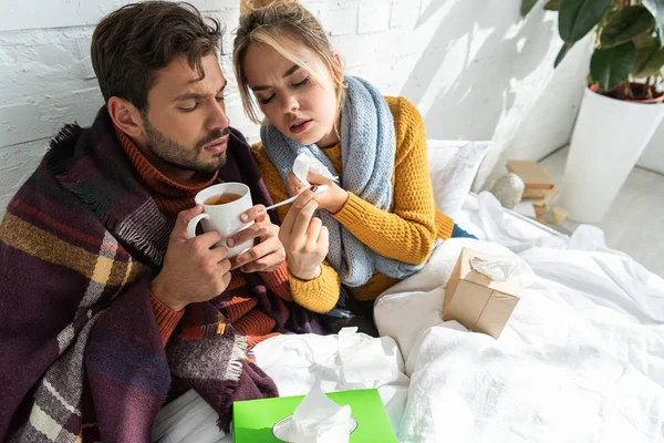Couple malade avec fièvre tenant thermomètre, boisson chaude et serviettes au lit — Photo de stock