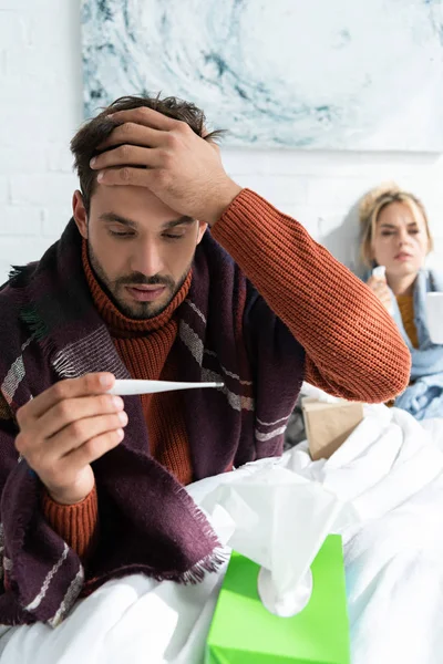 Больной мужчина с лихорадкой держит термометр в спальне с женщиной позади — стоковое фото
