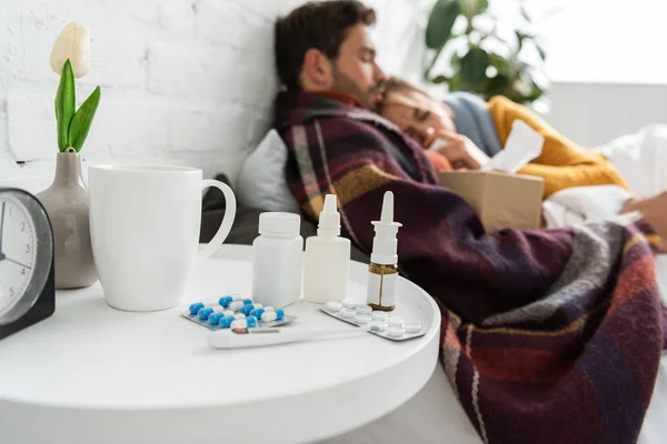 Selektive Fokussierung von Kranken im Bett mit Thermometer, Heißgetränk und Nasenspray und Tabletten auf dem Tisch — Stockfoto