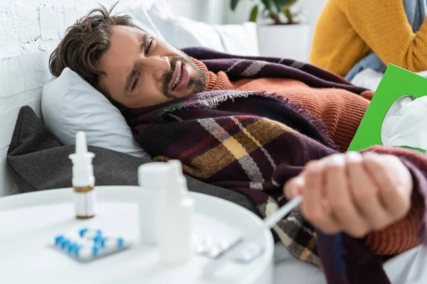 Hombre enfermo en la cama con pastillas, servilletas y aerosol nasal en el dormitorio con la mujer - foto de stock
