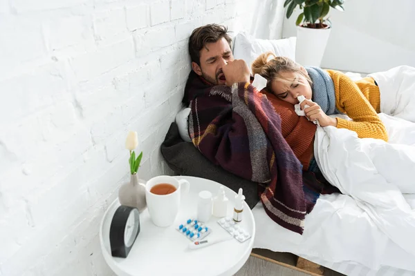 Хвора пара тримає серветки в спальні з термометром, таблетками, гарячим напоєм і носовим спреєм — стокове фото
