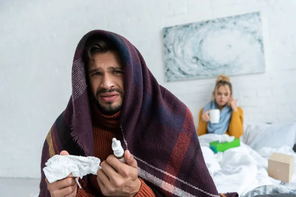 Uomo malato con spray nasale e tovagliolo in camera da letto con donna dietro — Foto stock