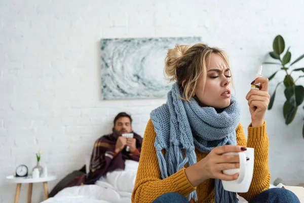 Sick woman holding nasal spray and hot drink in bedroom with man behind — Stock Photo