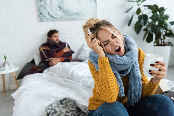 Mulher doente com dor de cabeça segurando bebida quente e spray nasal no quarto com o homem atrás — Fotografia de Stock