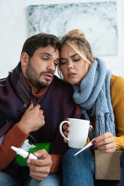 Sick couple with fever holding thermometer, hot drink and nasal spray — Stock Photo