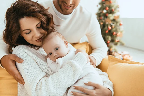 Cropped view of smiling man hugging wife sitting on sofa and holding infant — Stock Photo