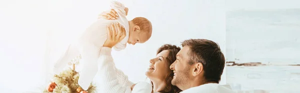 Tiro panorámico de hombre sonriente de pie cerca de esposa sosteniendo lindo bebé por encima de la cabeza - foto de stock