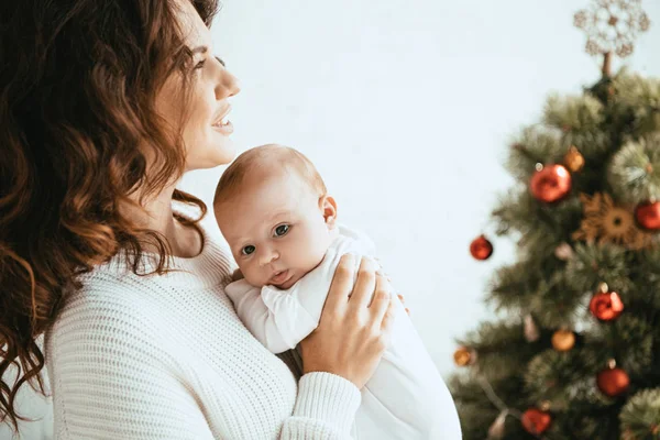Feliz madre sosteniendo adorable bebé mientras está de pie cerca del árbol de Navidad - foto de stock