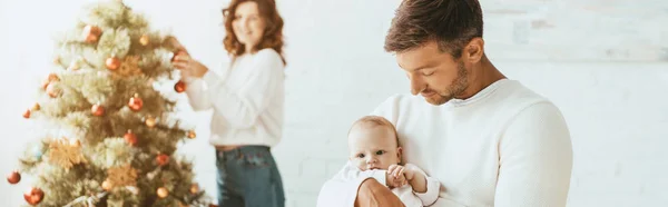 Tiro panorâmico de mulher decorando árvore de Natal e olhando para o marido feliz segurando bebê — Fotografia de Stock