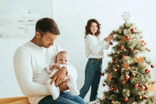 Selective focus of woman decorating christmas tree and looking at happy husband holding infant — Stock Photo