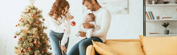 Ausgeschnittene Ansicht der Mutter zeigt Weihnachtskugel dem kleinen Kind, das auf den Händen des Vaters in der Nähe des geschmückten Weihnachtsbaums sitzt — Stockfoto