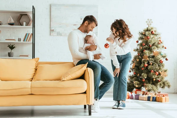 Attraktive Frau zeigt Weihnachtskugel einem kleinen Kind, das auf Väterhänden in der Nähe des geschmückten Weihnachtsbaums sitzt — Stockfoto