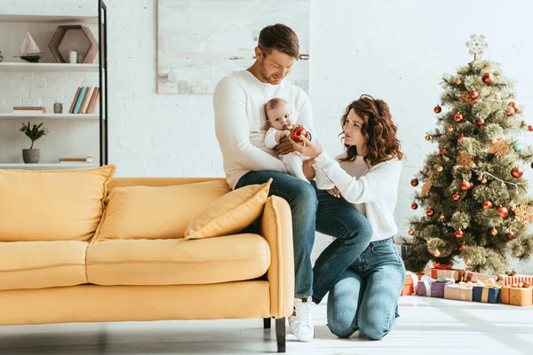 Attrayant femme donnant boule de Noël à bébé assis sur pères mains près décoré arbre de Noël — Photo de stock