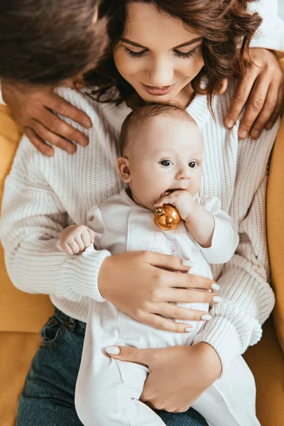 Visão cortada do homem olhando para o bebê adorável segurando bola de Natal enquanto sentado em mãos mães — Fotografia de Stock