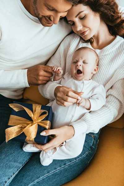 Mujer feliz sosteniendo caja de regalo mientras está sentado con el bebé adorable cerca del marido - foto de stock