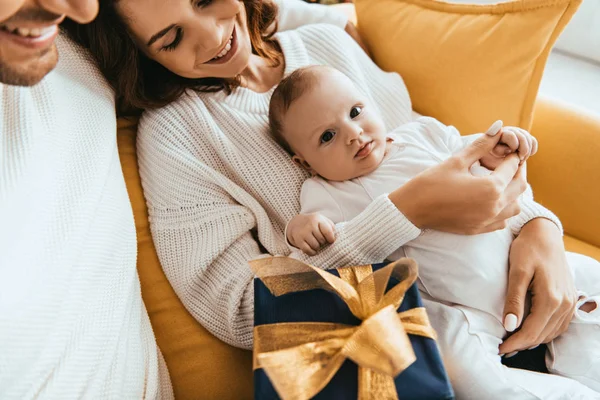 Vista recortada de hombre sonriente sentado cerca de esposa feliz sosteniendo adorable bebé cerca de caja de regalo con cinta de oro - foto de stock