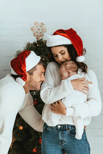 Attraktive Frau mit Weihnachtsmannmütze mit entzückendem Baby in der Nähe eines glücklichen Ehemanns — Stockfoto