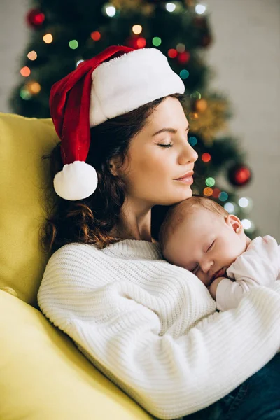 Junge Frau mit Weihnachtsmannmütze sitzt mit geschlossenen Augen und hält schlafendes Baby — Stockfoto