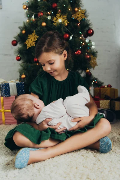 Adorable enfant tenant mignon petit sisiter tout en étant assis près de l'arbre de Noël et boîtes-cadeaux — Photo de stock