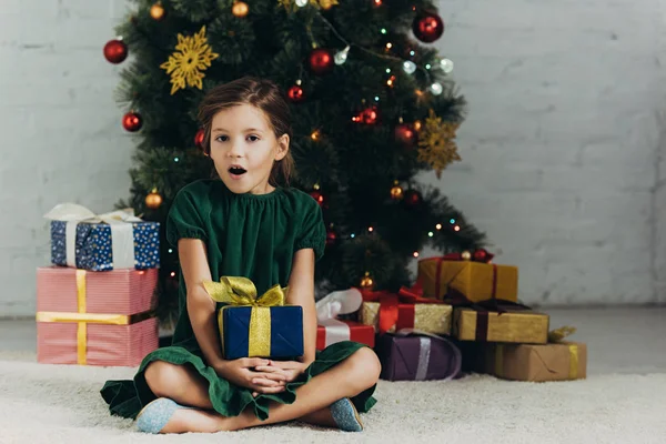 Удивленный ребенок сидит на полу возле рождественской елки, держит подарочную коробку и смотрит в камеру — стоковое фото